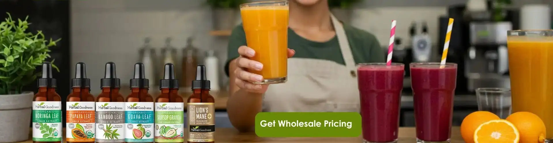 A woman in a green shirt and beige apron holds up a tall glass of bright orange juice, with several bottles of herbal tinctures lined up on the counter beside her.